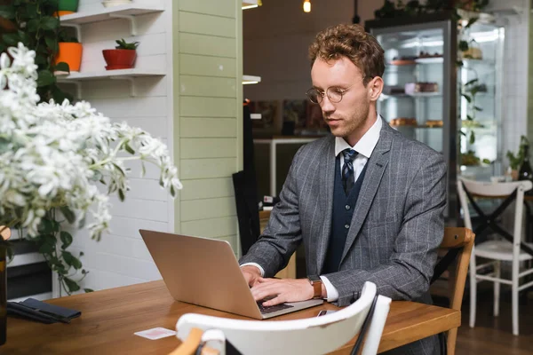 Кучерявий бізнесмен у костюмі та окулярах, використовуючи ноутбук у кафе — стокове фото