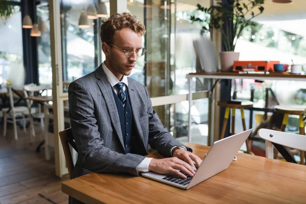 Homem de negócios encaracolado em terno e óculos digitando no laptop no café — Fotografia de Stock