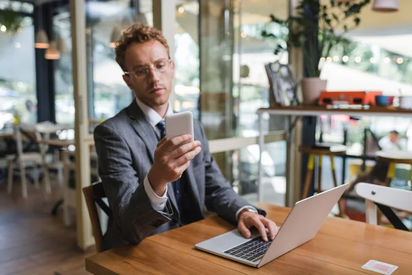 Curly businessman in suit and glasses using smartphone near laptop in cafe — Stock Photo