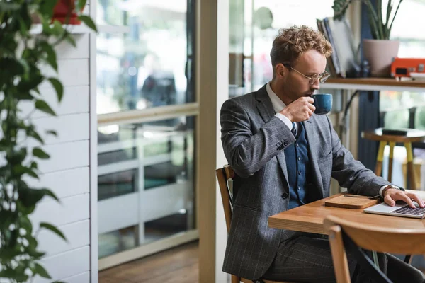 Curly businessman in formal wear and glasses using laptop and drinking coffee in cafe — Stock Photo