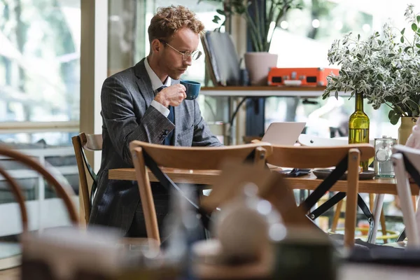 Homem de negócios encaracolado em desgaste formal e óculos usando laptop e segurando xícara de café no café — Fotografia de Stock