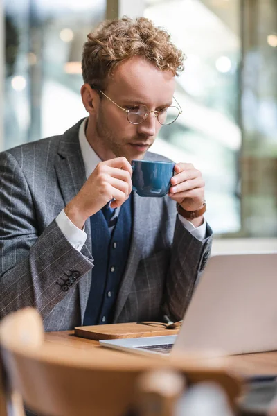 Кудрявый бизнесмен в очках с помощью ноутбука и с чашкой кофе в кафе — стоковое фото