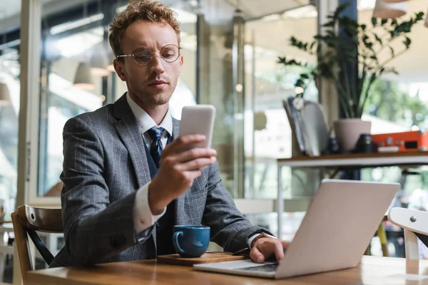 Homem de negócios encaracolado em óculos usando telefone celular perto de laptop e xícara com café no café — Fotografia de Stock