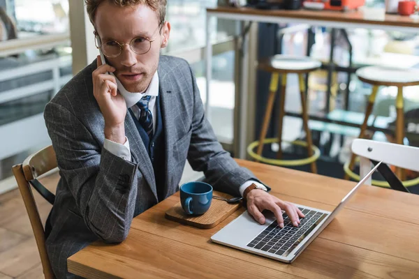 Hombre de negocios rizado en gafas hablando en el teléfono móvil cerca de la computadora portátil y la taza con café en la cafetería - foto de stock