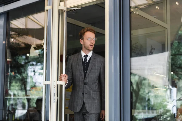 Homme d'affaires bouclé dans des lunettes ouvrant la porte tout en quittant le café — Photo de stock