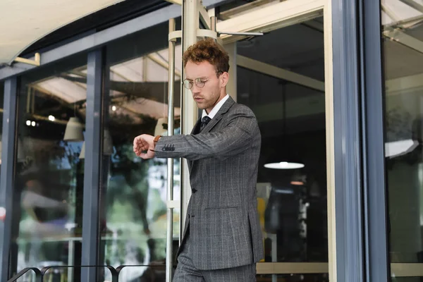 Curly businessman in glasses looking at wristwatch while leaving cafe — Stock Photo