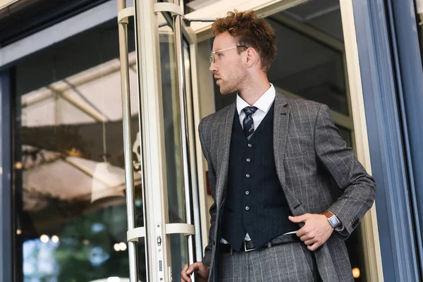 Uomo d'affari riccio con gli occhiali aprendo la porta e guardando lontano mentre lascia il caffè — Foto stock