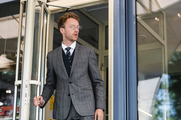 Hombre de negocios rizado en gafas y elegante traje abriendo la puerta mientras sale de la cafetería - foto de stock
