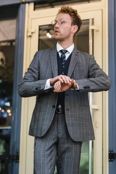 Homem de negócios encaracolado em terno elegante ajustando relógio de pulso perto da entrada do café — Fotografia de Stock