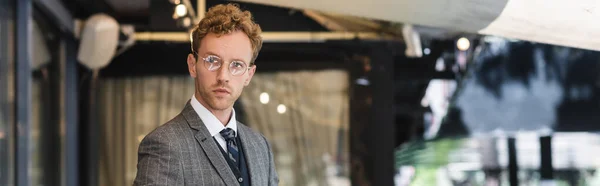 Lockiger Geschäftsmann mit Brille und edlem Anzug in der Nähe des Caféeingangs, Banner — Stockfoto