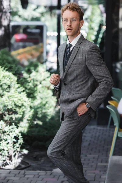 Jovem empresário em óculos e terno elegante de pé com a mão no bolso perto da entrada do café — Fotografia de Stock