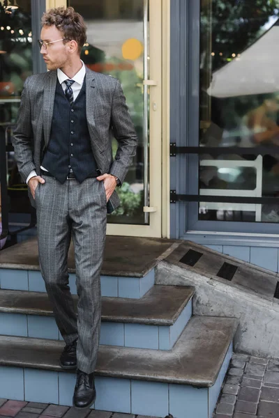 Curly businessman in glasses and classy suit walking with hands in pockets near cafe entrance — Stock Photo