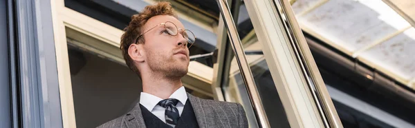 Vista basso angolo di uomo d'affari riccio in occhiali e vestito di classe porta di apertura lasciando caffè, banner — Foto stock