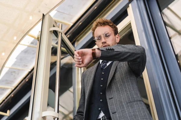 Low angle view of curly businessman in glasses looking at wristwatch while leaving cafe — Stock Photo