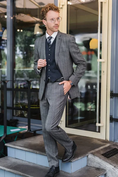 Hombre de negocios rizado en gafas y traje elegante caminando con la mano en el bolsillo cerca de la entrada de la cafetería - foto de stock