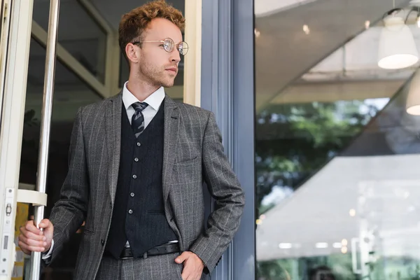 Curly businessman in glasses and classy suit standing with hand in pocket near cafe entrance — Stock Photo