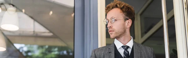 Hombre de negocios en gafas y traje elegante mirando hacia otro lado cerca de la entrada de la cafetería, pancarta - foto de stock