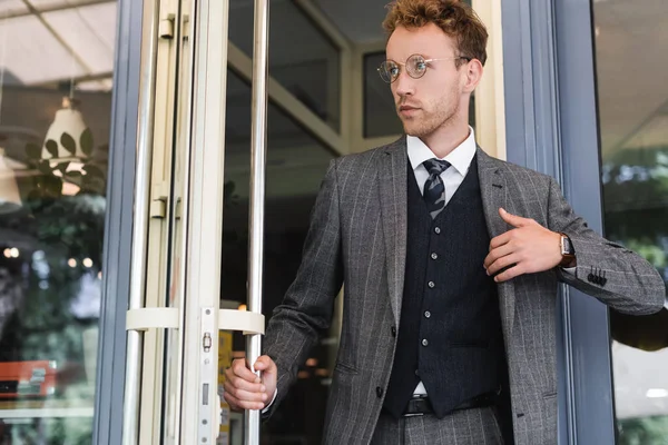 Young and curly businessman in glasses opening door while leaving cafe — Stock Photo