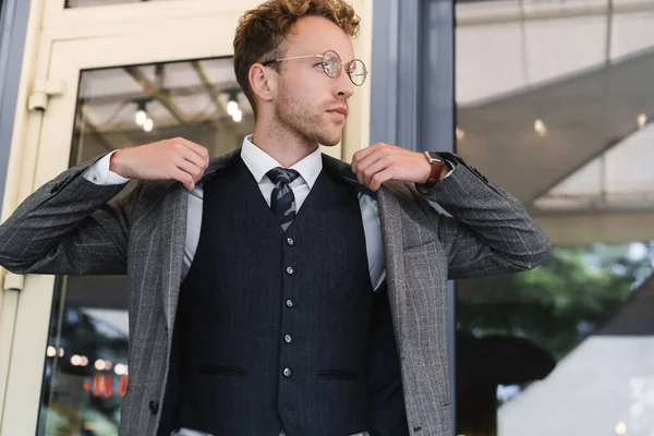 Homem de negócios encaracolado em óculos e terno elegante vestindo blazer perto da entrada do café — Fotografia de Stock