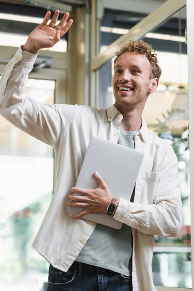 Positive freelancer holding laptop and waving hand while entering cafe — Stock Photo