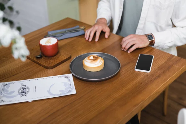 Vista recortada del hombre cerca de tarta de limón, capuchino, menú y teléfono inteligente con pantalla en blanco en la mesa - foto de stock