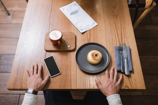 High angle view of man near lemon tart, cappuccino and menu near smartphone with blank screen on table — Stock Photo