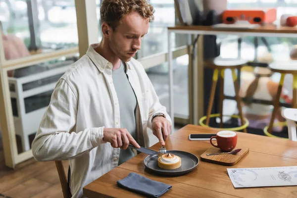 Curly man holding cutlery and looking at lemon tart near cappuccino and smartphone with blank screen on table — Stock Photo