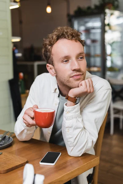 Nachdenklicher Mann hält Tasse Cappuccino in der Hand und schaut im Café weg — Stockfoto