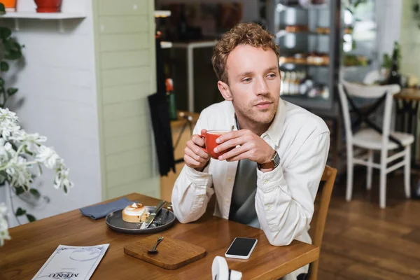 Bouclé homme tenant tasse de cappuccino près gadget et tarte au citron tout en regardant loin dans le café — Photo de stock