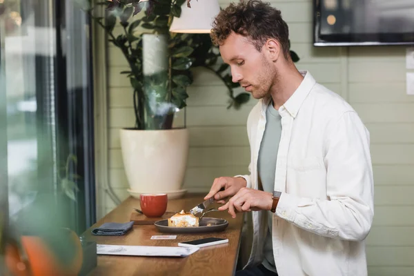 Side view of curly man holding cutlery near lemon tart on plate, smartphone and cup in cafe — Stock Photo