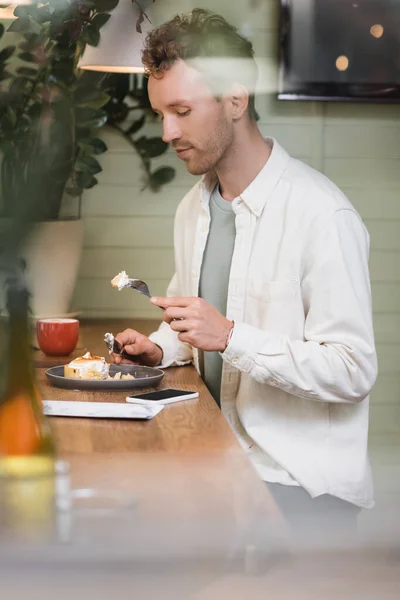 Кудрявый человек держит вилку с кусочком лимонного пирога рядом со смартфоном и чашкой в кафе — стоковое фото
