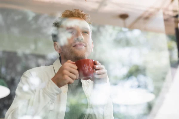 Homme bouclé tenant tasse de café derrière la fenêtre floue dans le café — Photo de stock