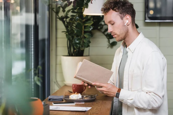 Hombre rizado en auriculares inalámbricos libro de lectura en la cafetería - foto de stock