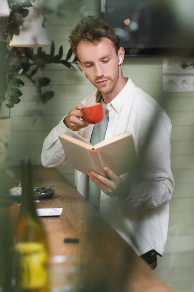 Curly man in wireless earphones reading book while holding cup in blurred cafe — Stock Photo