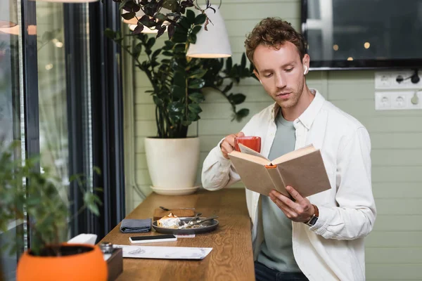 Young man in wireless earphones reading book while holding cup of coffee in blurred cafe — Stock Photo
