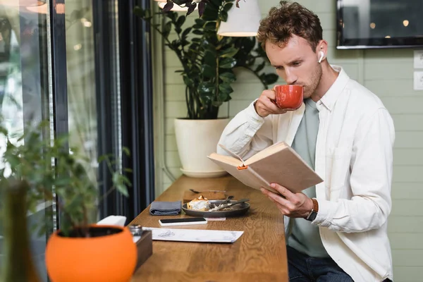 Joven hombre rizado en auriculares inalámbricos libro de lectura mientras bebe café en café borroso - foto de stock
