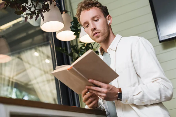 Low angle view of young curly man in wireless earphones reading book in cafe — Stock Photo