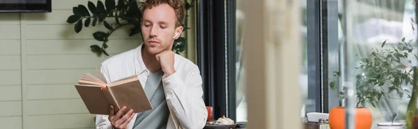 Young curly man in wireless earphones reading book in cafe, banner — Stock Photo