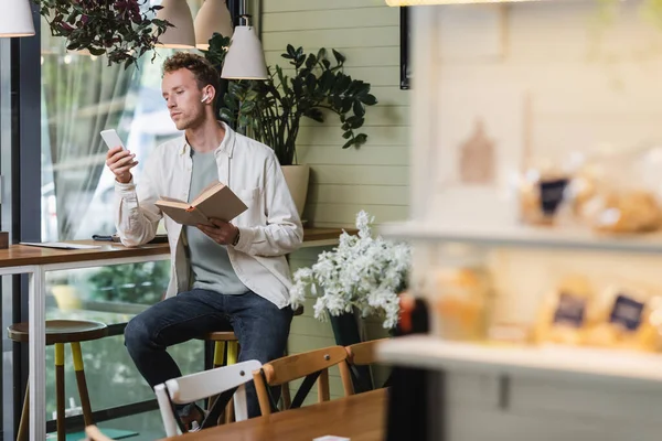 Hombre rizado en auriculares inalámbricos con teléfono inteligente y libro de celebración en la cafetería - foto de stock