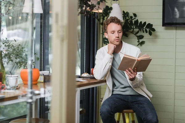 Сосредоточенный человек в беспроводных наушниках чтение книги в кафе — стоковое фото