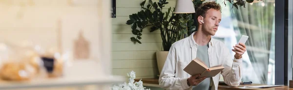 Hombre rizado en auriculares inalámbricos con teléfono inteligente y libro de celebración en la cafetería, pancarta — Stock Photo