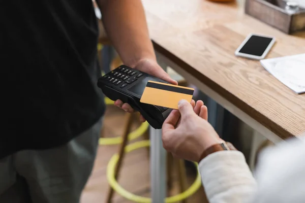 Vista cortada de garçom segurando terminal de pagamento perto do cliente com cartão de crédito no café — Fotografia de Stock