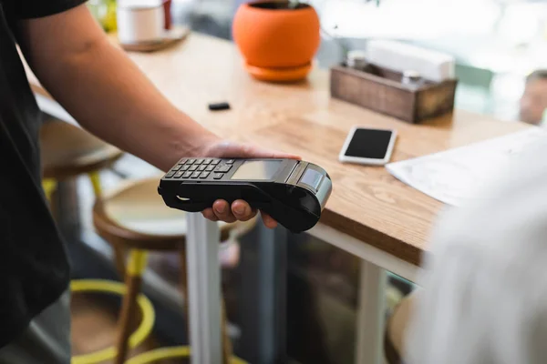 Ausgeschnittene Ansicht des Kellners mit Zahlungsterminal in der Nähe eines verschwommenen Kunden im Café — Stockfoto