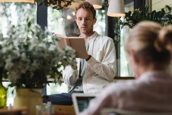 Curly man in wireless earphones reading book near blurred customer in cafe — Stock Photo