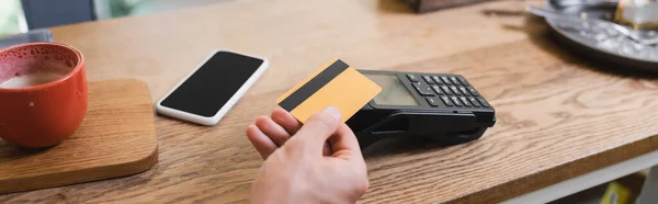 Vista recortada del hombre que paga con tarjeta de crédito cerca de teléfono inteligente en la cafetería, pancarta - foto de stock