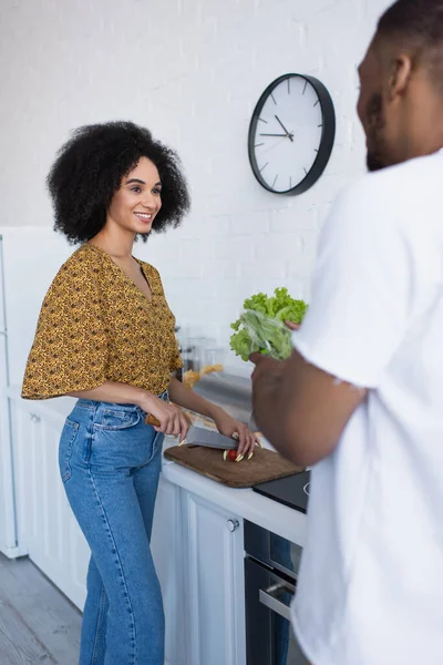 Sonriente mujer afroamericana cortando tomate cherry cerca de novio borroso en cocina - foto de stock