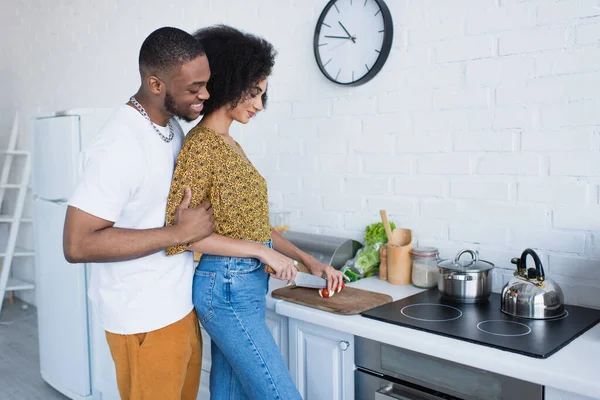 Souriant homme afro-américain étreignant petite amie coupe salade dans la cuisine — Photo de stock