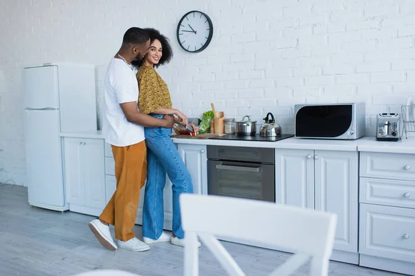 Afro-américain homme étreignant petite amie souriante coupe salade fraîche dans la cuisine — Photo de stock