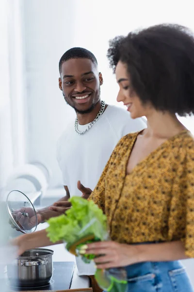 Sonriente hombre afroamericano mirando a su novia con lechuga en primer plano borroso - foto de stock