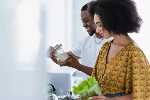 Vue latérale d'un homme afro-américain tenant un pot avec du riz près d'une petite amie floue dans la cuisine — Photo de stock